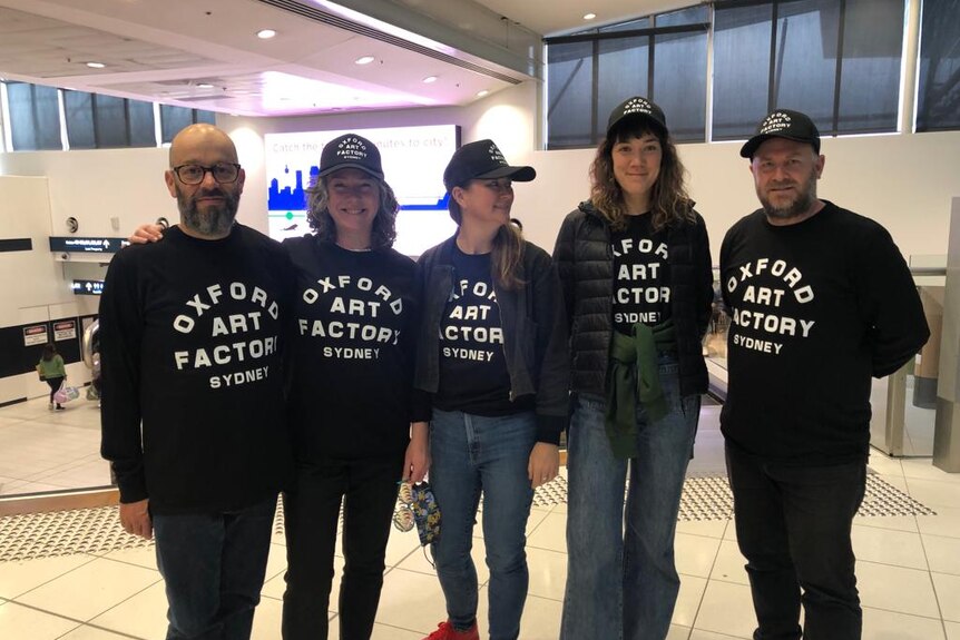Five members of Frente! at the airport all wearing matching Ocxford Art Factory shirts and caps