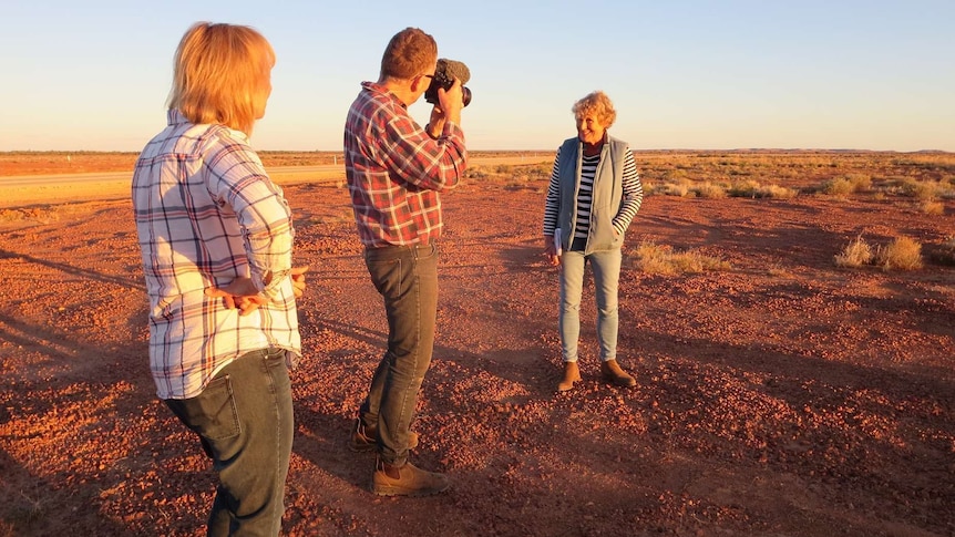 Cameraman Campbell Miller films Heather Ewart talking to camera as producer Lisa Whitehead watches, with red desert backdrop.