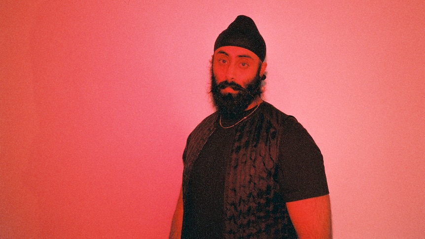 Yung Singh wears a black turban & has a black beard and moustache. He wears a black tee with vest & stands in bright red light.
