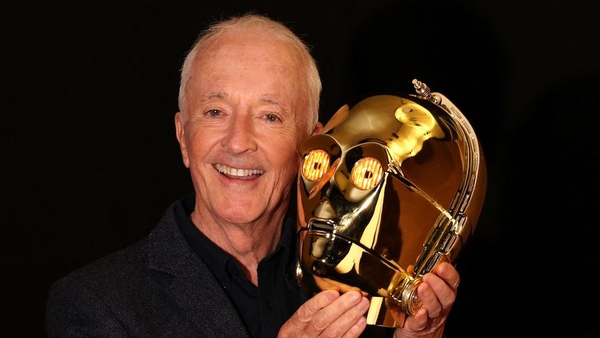 Anthony Daniels smiles holding up a gold light-up robotic looking head.