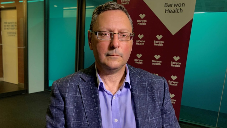 A man wearing a blue striped suit, blue shirt and glasses stares at the camera, behind him a Barwon Health banner.