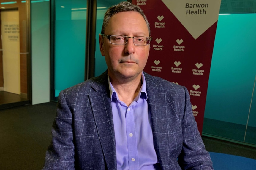 A man wearing a blue striped suit, blue shirt and glasses stares at the camera, behind him a Barwon Health banner.