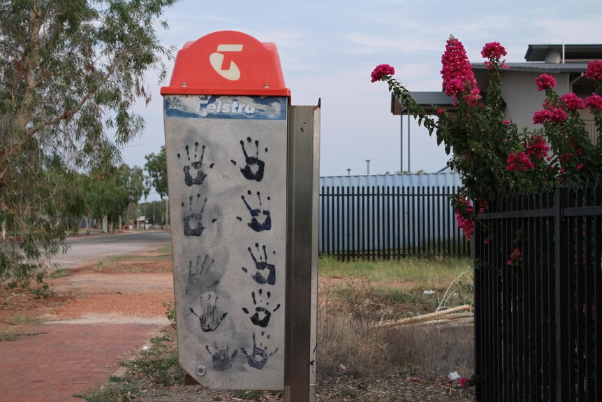 Un teléfono público de Telstra cubierto de huellas de manos negras, junto a una carretera y un sendero de tierra roja.