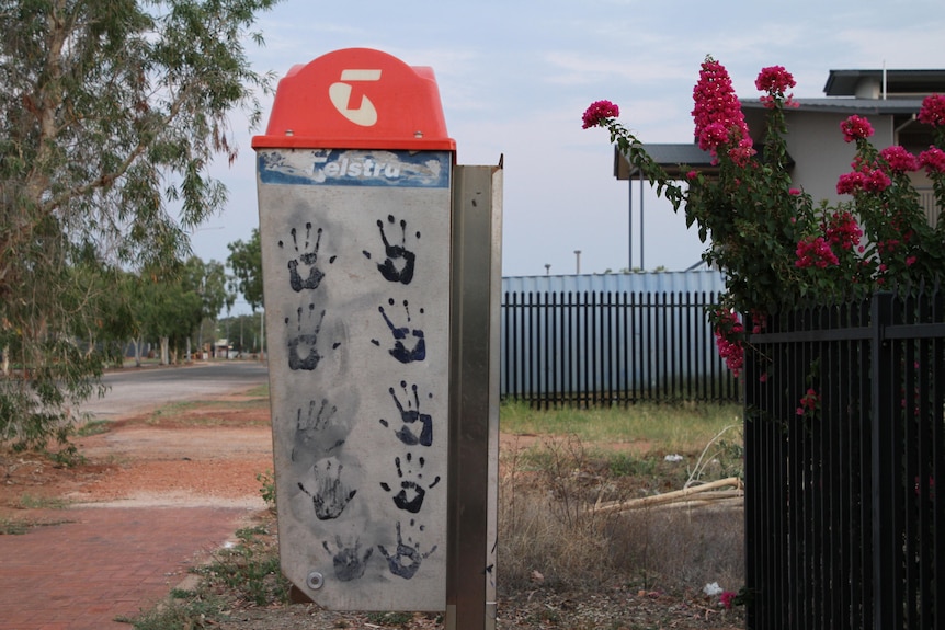 A Telstra pay phone covered in black pain handprints, next to a road and path of red earth.