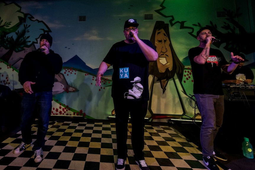 Three men wearing dark coloured clothes holding microphones and standing on a black and white platform.