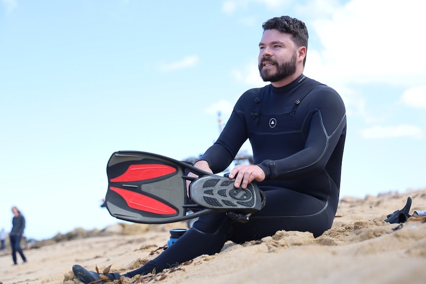 A man with a beard in a wetsuit sits in the sand pulling on a flipper.