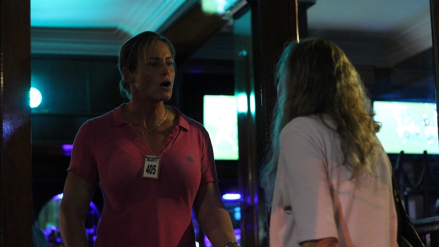 A muscular woman on the door of a nightclub talks heatedly to another woman facing away from the camera.