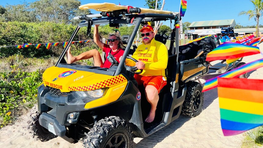 Two male lifesavers in a beach buggy driving down a beach access road lined with rainbow pride flags. 