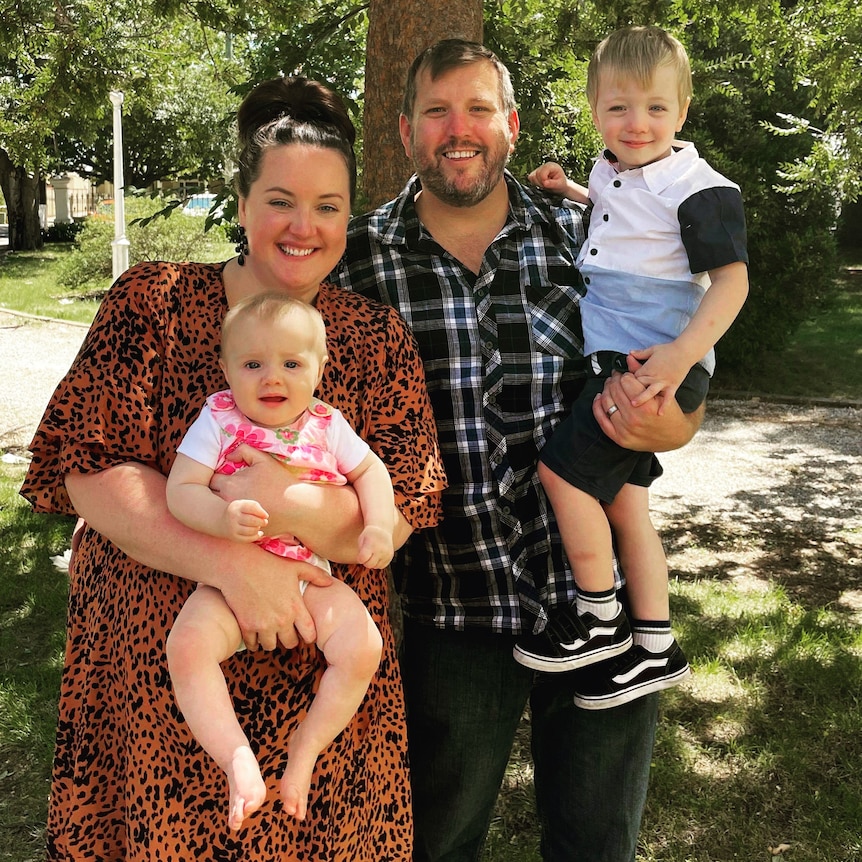 A family of four stand in front of a tree, smiling at the camera.