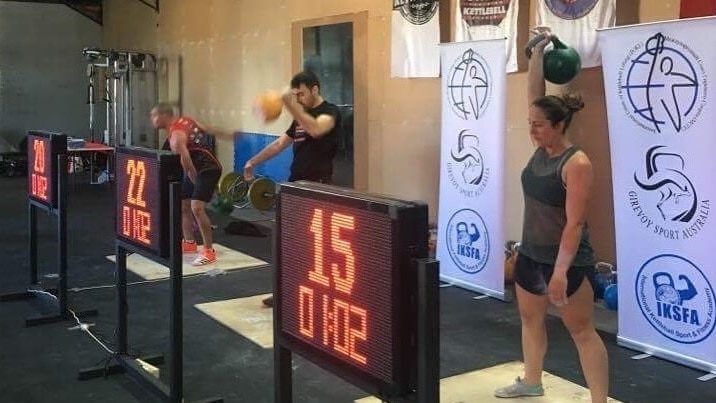Three people are lifting kettlebells with one hand in front of electronic signs