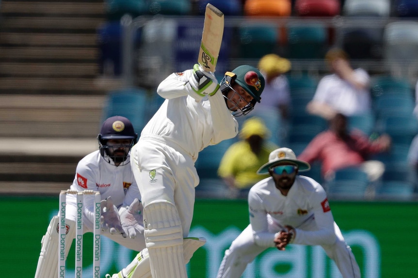 A man swings his bat and hits a red cricket ball towards the camera as two men crouching behind him look on