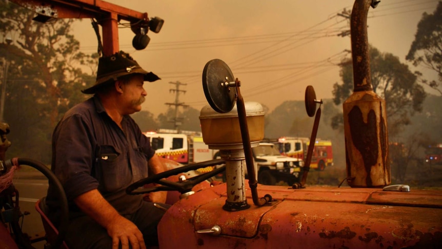 A farmer sits on a tractor surrounded by a red haze from a nearby bushfire. Fire trucks in the background.
