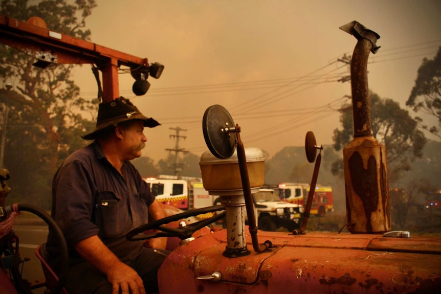 A farmer sits on a tractor surrounded by a red haze from a nearby bushfire. Fire trucks in the background.