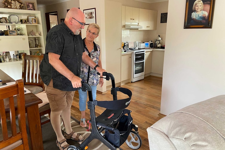 Man stands holding a walking frame with a woman next to him inside a house