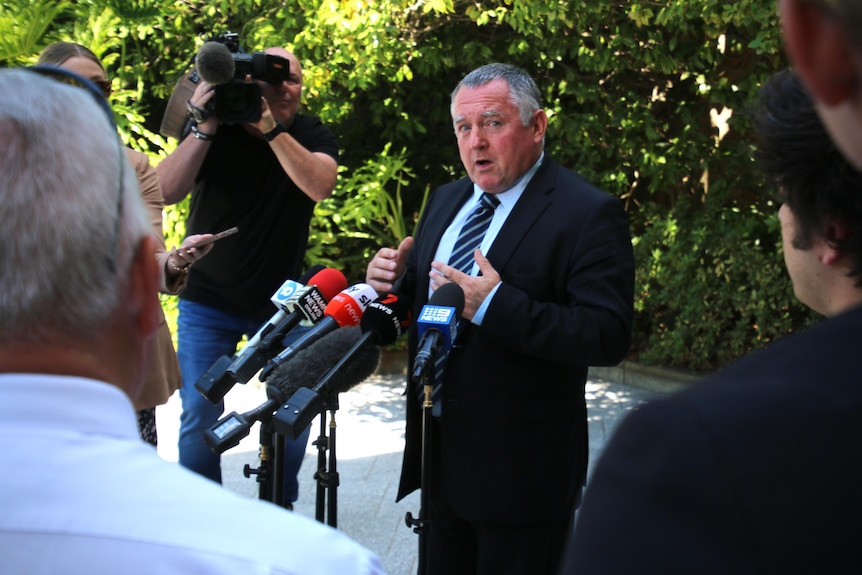 A man wearing a black suit stands at a press conference