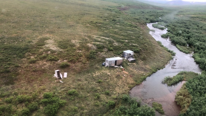 Three ramshackle buildings in the wilderness near a river.