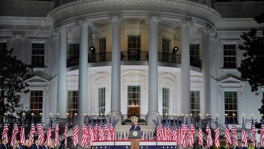 A man in a suit stands on a podium in front of a White house with flags in the background and a crowd in front.