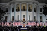 A man in a suit stands on a podium in front of a White house with flags in the background and a crowd in front.