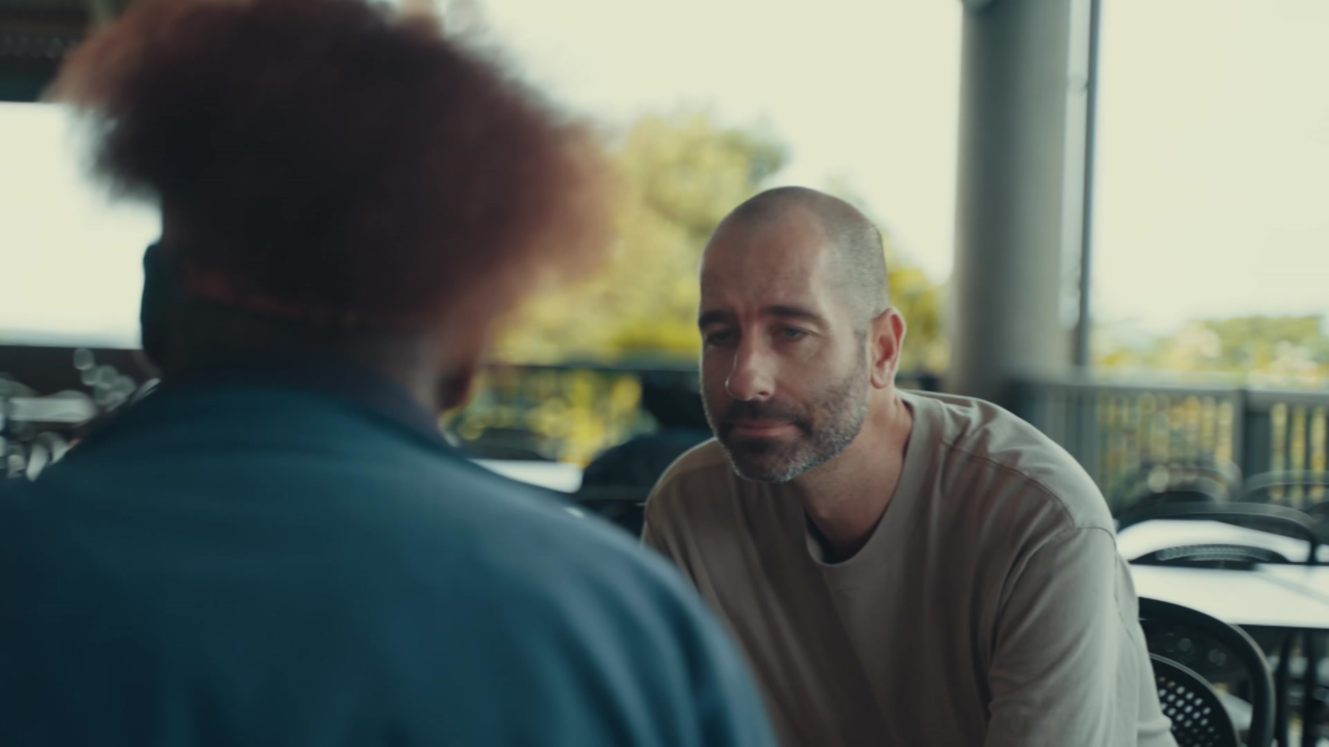 A man in a beige T-shirt talking to another person.