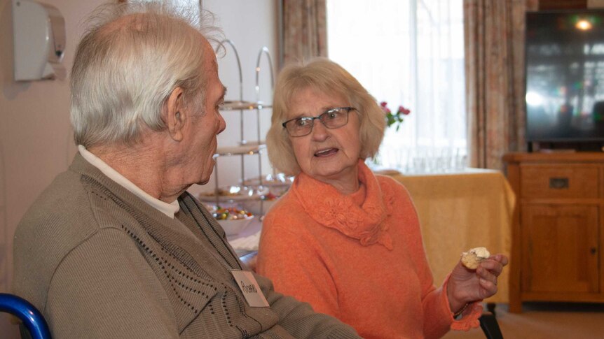 An ageing man and woman sit together inside an aged care home. They are looking at each other and talking.
