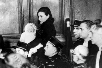 Black and white photo of a standing in court, holding a baby, while flanked by police.