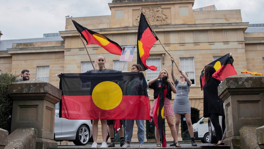 A group of women wave Aboriginal flags in front of a stone building