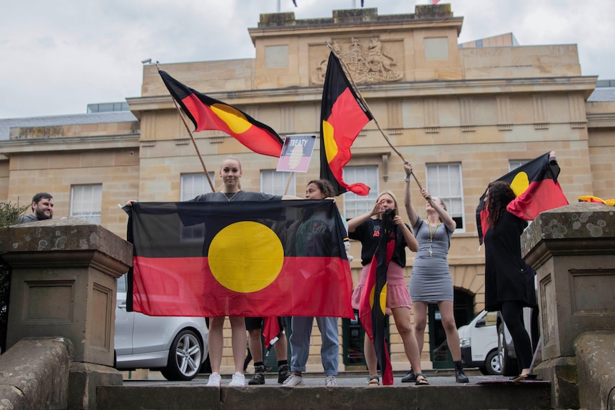 A group of women wave Aboriginal flags in front of a stone building