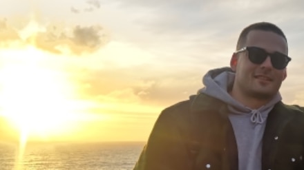 A photo of a young man in a grey hoodie and black jacket smiling in front of a sunset over water.