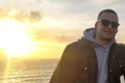 A photo of a young man in a grey hoodie and black jacket smiling in front of a sunset over water.