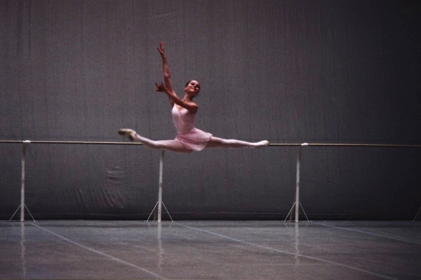 Girl in pink ballet costume does the splits in the air in a dance studio.