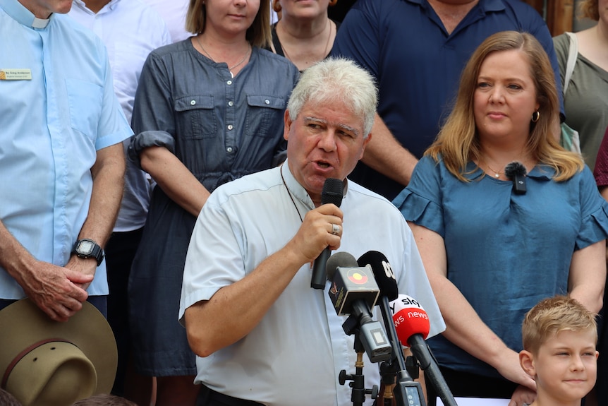 A grey-haired man and a brown-haired woman hold microphones on the steps of a church with a crowd behind them