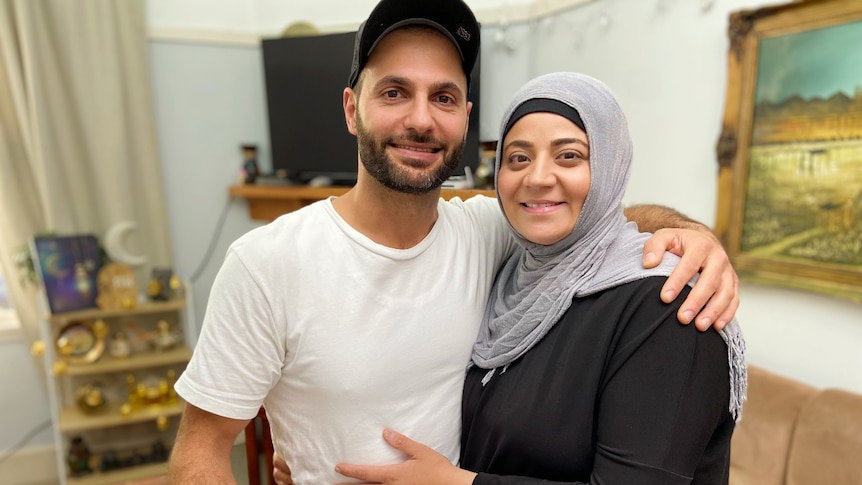 A bearded man wearing a hat embarces a woman in dark clothing wearing a grey head scarf.