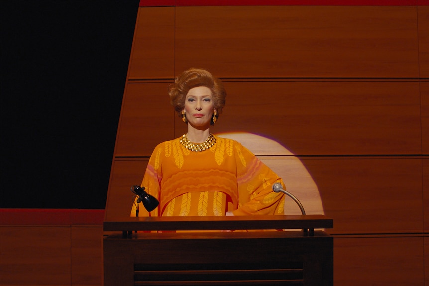 50-something woman with bouffant orange hair and bright makeup spotlit at podium, poker faced.