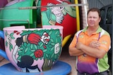middle aged man standing next to a tea cup ride wearing an orange, purple and geen shirt with his arms folded.