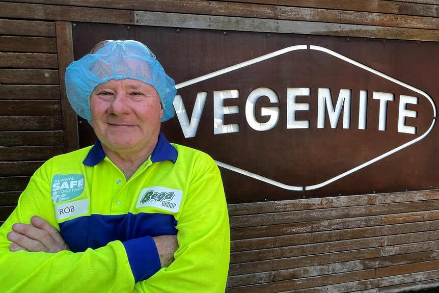 Man in fluorescent yellow jumper and hair net smiles at camera in front of wrought iron vegemite sign 