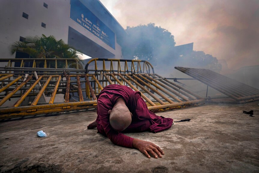 A Buddhist monk in a red robe lies on the ground outside police headquarters, with a cloud of tear gas in the background. 