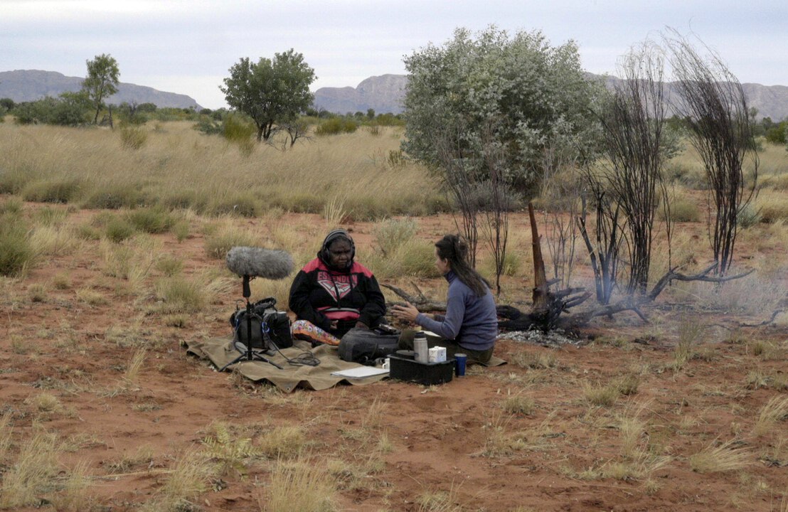 An Aboriginal woman sits with a researcher who has sound recording equipment in a rural landscape.