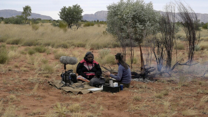 An Aboriginal woman sits with a researcher who has sound recording equipment in a rural landscape.