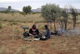 An Aboriginal woman sits with a researcher who has sound recording equipment in a rural landscape.