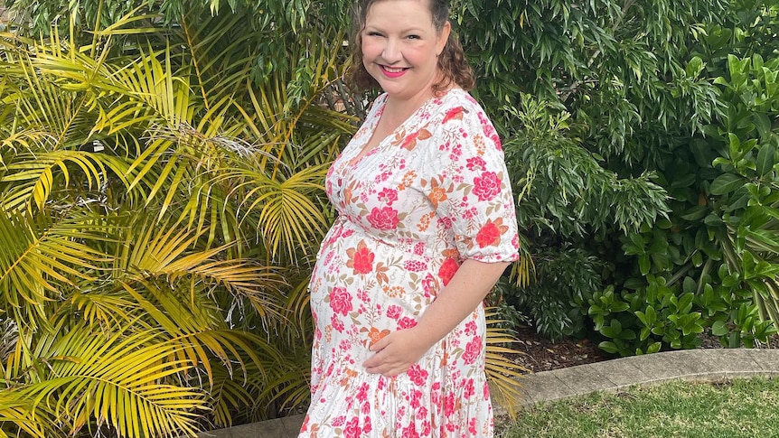 A woman standing in the backyard in a summery dress holds her hands on a small baby bump.