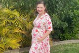 A woman standing in the backyard in a summery dress holds her hands on a small baby bump.