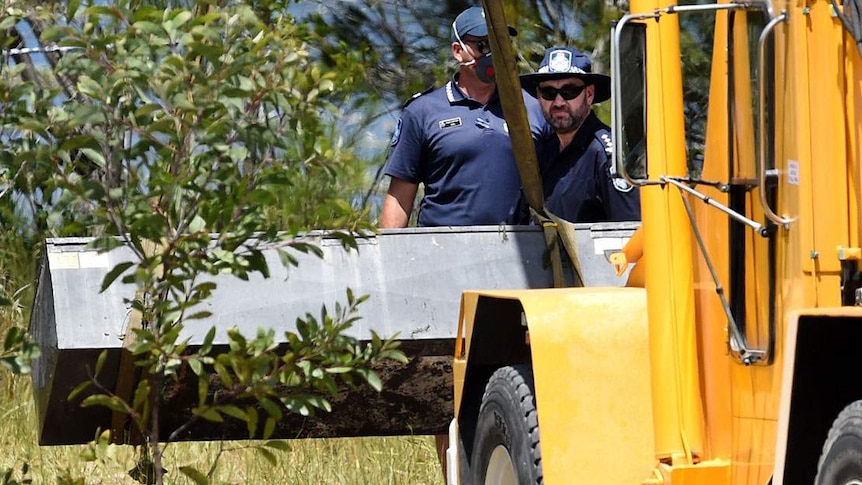 Two policemen watch as a yellow crane truck lifts a black box in grassland