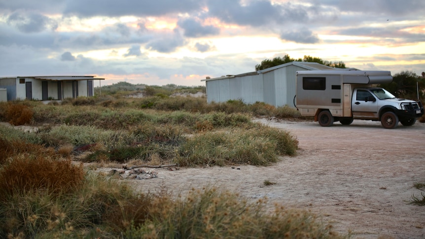 A sandy camp site with an old campfire visible and tin shacks and a vehicle in the background.
