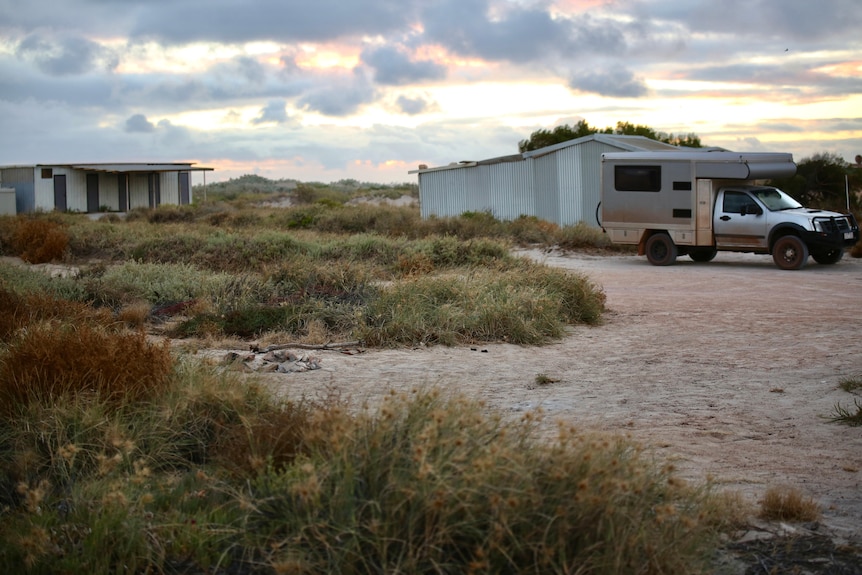 A sandy camp site with an old campfire visible and tin shacks and a vehicle in the background.