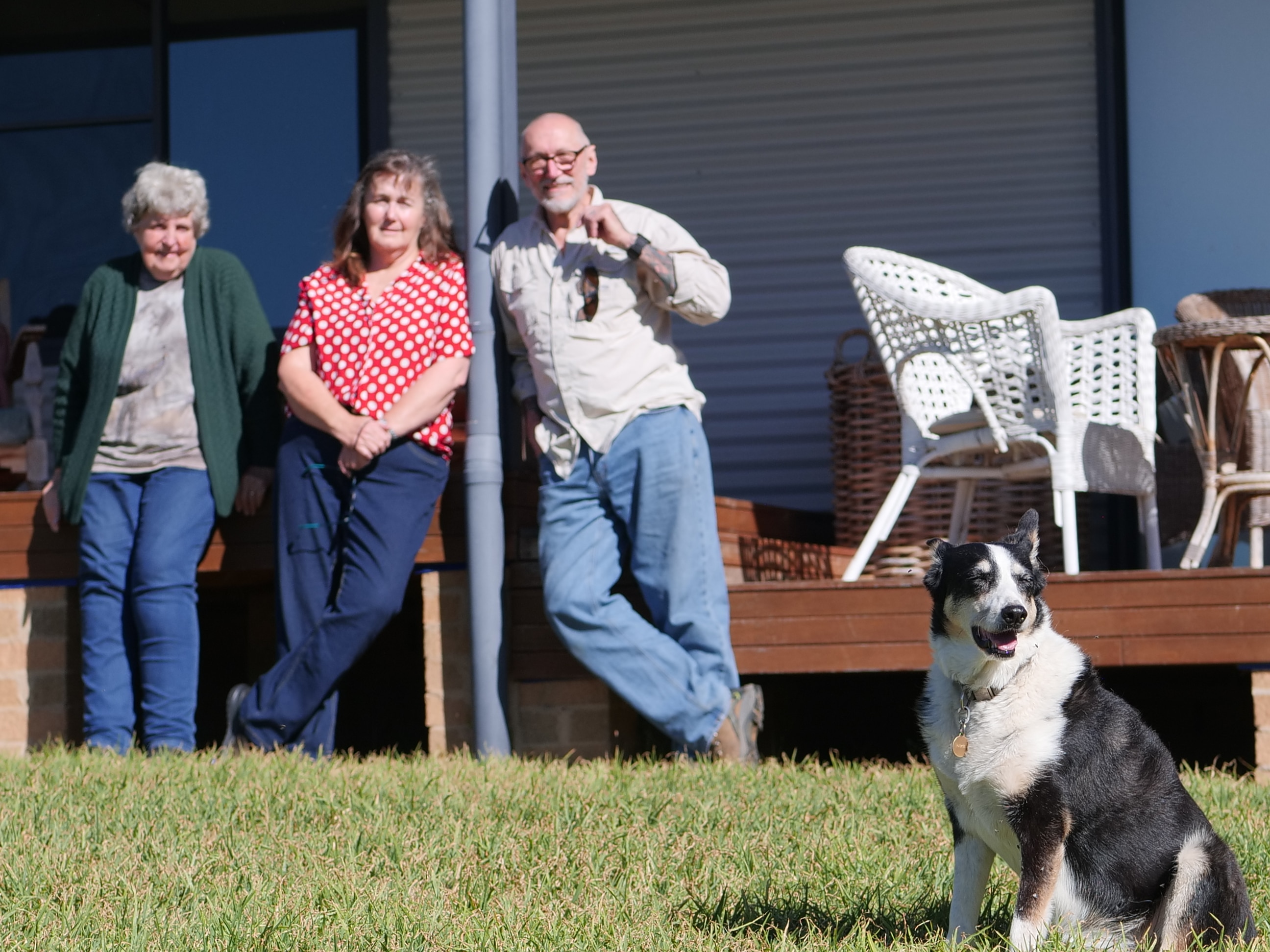 Three people in background leaning against deck, dog in foreground. 