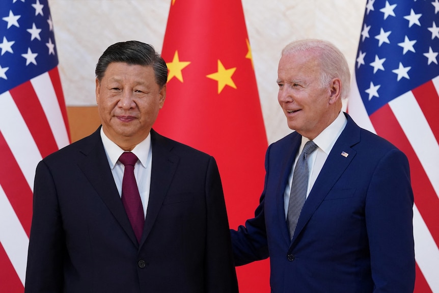 US President Joe Biden smiles and holds a hand behind Chinese President Xi Jinping in front of US and Chinese flags