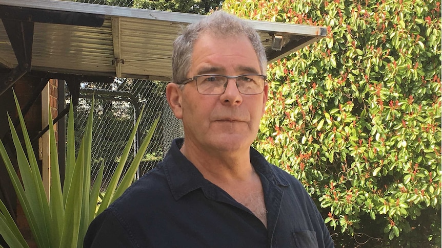 A man with grey hair, a blue button-up shirt and a serious expression pictured in a garden.