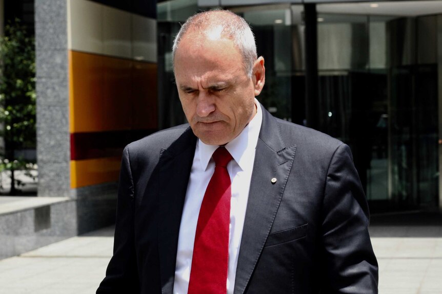 NAB chairman Ken Henry, wearing a red tie with his suit, leaves the royal commission with his head down and brow furrowed.