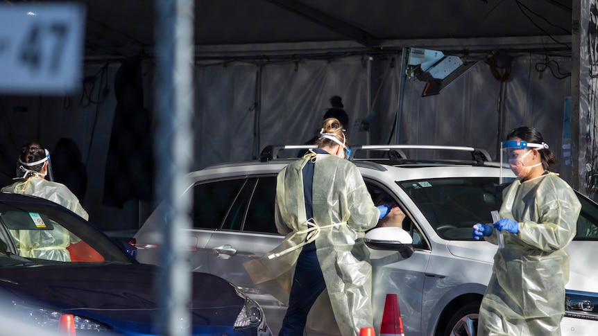 Three women in masks and gowns next to a car at a covid testing clinic.