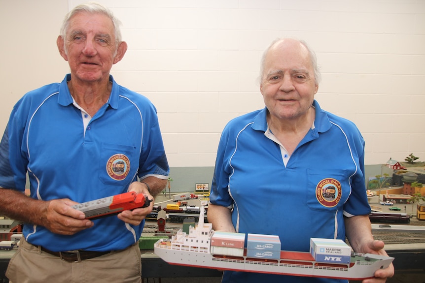 Two men in blue shirts standing side by side holding a model train and boat
